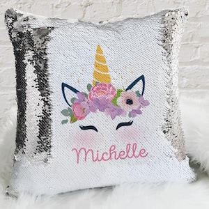 Puede incluir: Una almohada de lentejuelas blancas y plateadas con un diseño de unicornio. El unicornio tiene un cuerno amarillo, flores rosas y una cara rosa. El nombre "Michelle" está escrito en letras rosas debajo del unicornio.