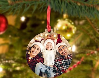 Op de afbeelding: Een ronde houten kerstbal met een foto van een gezin dat kerstmanmutsen draagt. De foto toont een vrouw, een kind en een man die lachen en op een witte ondergrond liggen met kerstverlichting.