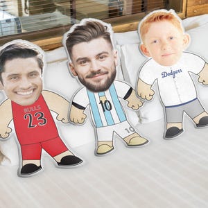 May include: Three custom face pillows with cartoon bodies. The pillow on the left is wearing a red basketball jersey with the number 23 and the word "BULLS". The middle pillow is wearing a light blue and white soccer jersey with the number 10. The pillow on the right is wearing a white baseball jersey with the word "Dodgers".
