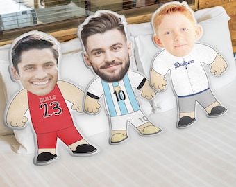 May include: Three custom face pillows with cartoon bodies. The pillow on the left is wearing a red basketball jersey with the number 23 and the word "BULLS". The middle pillow is wearing a light blue and white soccer jersey with the number 10. The pillow on the right is wearing a white baseball jersey with the word "Dodgers".