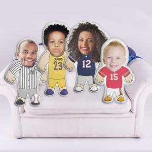 May include: Four custom-made pillows with faces printed on them. Each pillow is dressed in a different sports uniform. The pillows are sitting on a white couch.