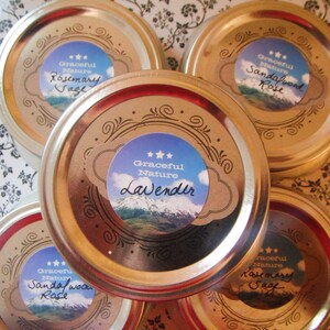 May include: An overhead shot of five jars with gold-colored lids. Each lid has a label with the words "Graceful Nature" and a scent name like "Rosemary Sage" or "Lavender." The labels feature a mountain scene.