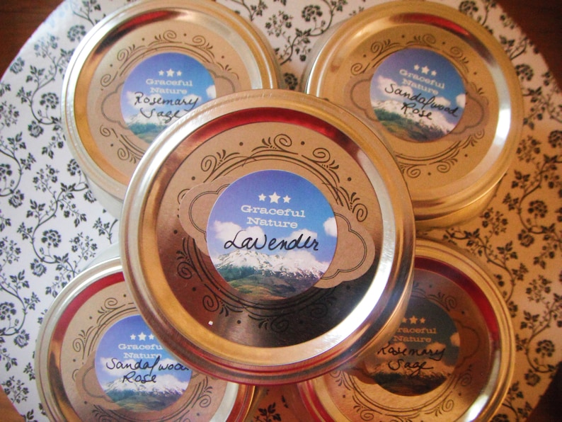 May include: An overhead shot of five jars with gold-colored lids. Each lid has a label with the words "Graceful Nature" and a scent name like "Rosemary Sage" or "Lavender." The labels feature a mountain scene.