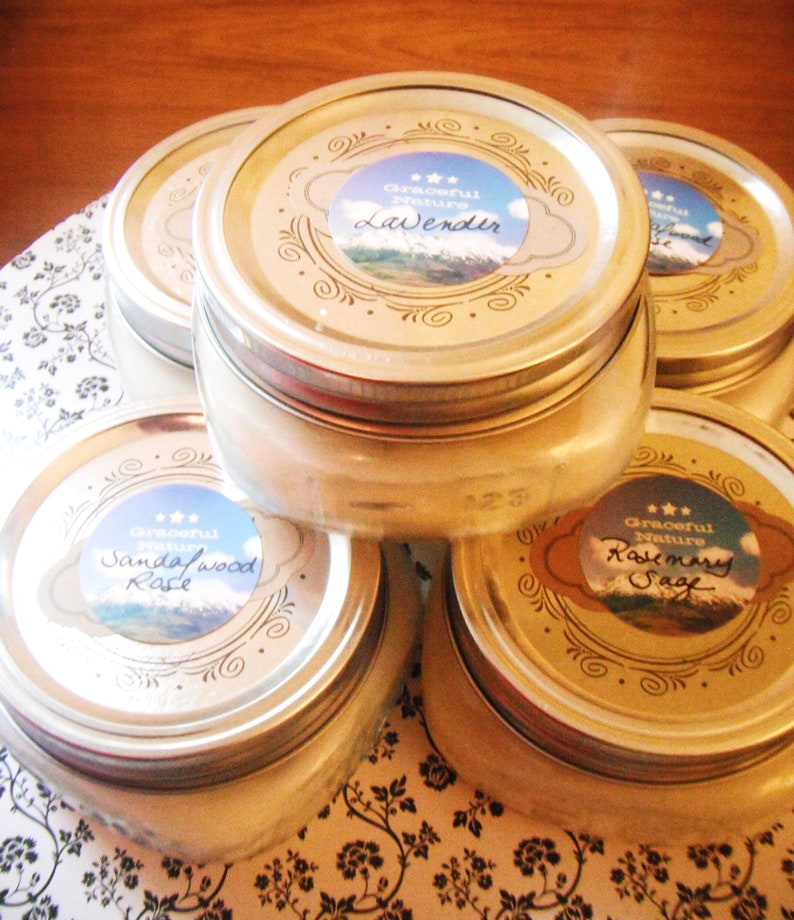 May include: Several glass jars with silver lids are stacked on a floral patterned surface. Each jar has a label with the words "Graceful Nature" and a scent name: Lavender, Sandalwood Rose, or Rosemary Sage. The jars are filled with a creamy substance.
