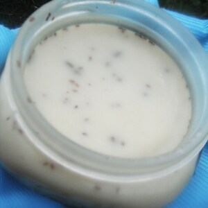 May include: A clear plastic jar filled with a creamy white substance speckled with small, dark particles. The jar has a wide opening and is sitting on a blue cloth. The contents appear to be a cosmetic product.