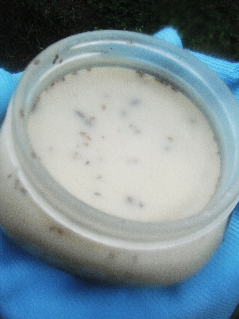 May include: A clear plastic jar filled with a creamy white substance speckled with small, dark particles. The jar has a wide opening and is sitting on a blue cloth. The contents appear to be a cosmetic product.