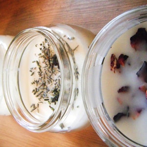 May include: Three glass jar candles with creamy white wax. One candle is topped with lavender sprigs, while another features dried rose petals. The jars sit on a wooden surface, offering a rustic aesthetic.