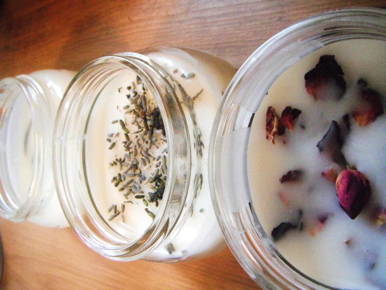 May include: Three glass jar candles with creamy white wax. One candle is topped with lavender sprigs, while another features dried rose petals. The jars sit on a wooden surface, offering a rustic aesthetic.