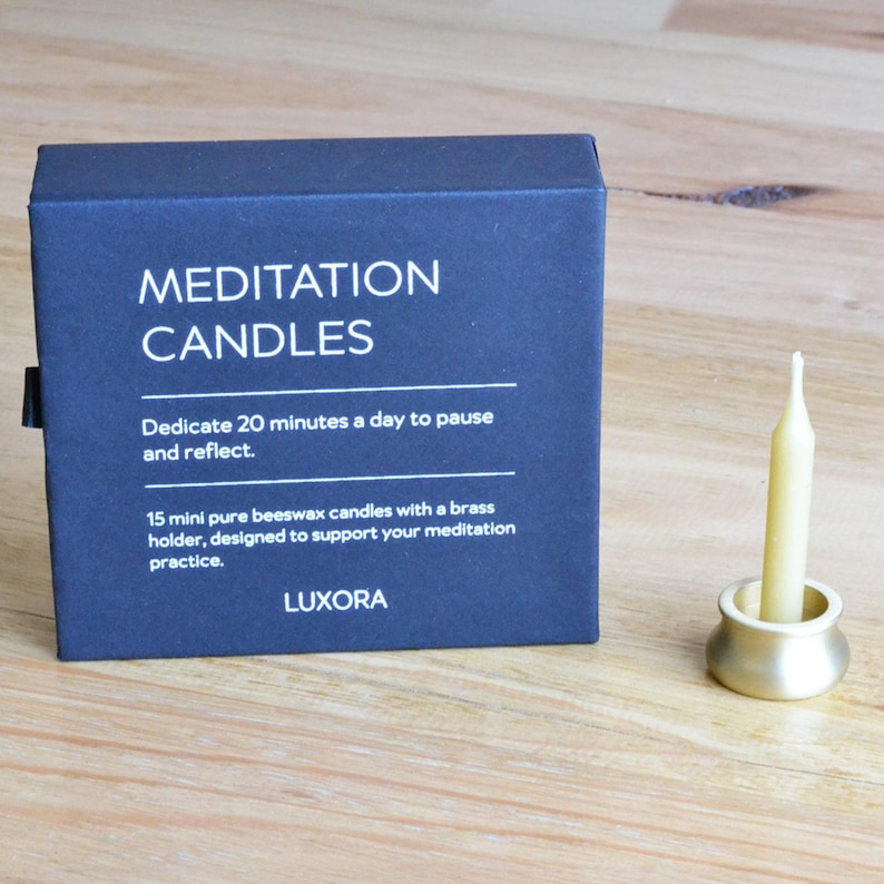 May include: A navy blue box with the words "MEDITATION CANDLES" in white. The box contains 15 mini beeswax candles with a brass holder. A lit candle sits in a brass holder. The text on the box says to dedicate 20 minutes a day to pause and reflect.