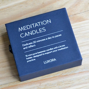 May include: A navy blue box of meditation candles. The box reads "MEDITATION CANDLES" and includes text about dedicating 20 minutes a day to pause and reflect. It also mentions 15 mini beeswax candles with a brass holder.