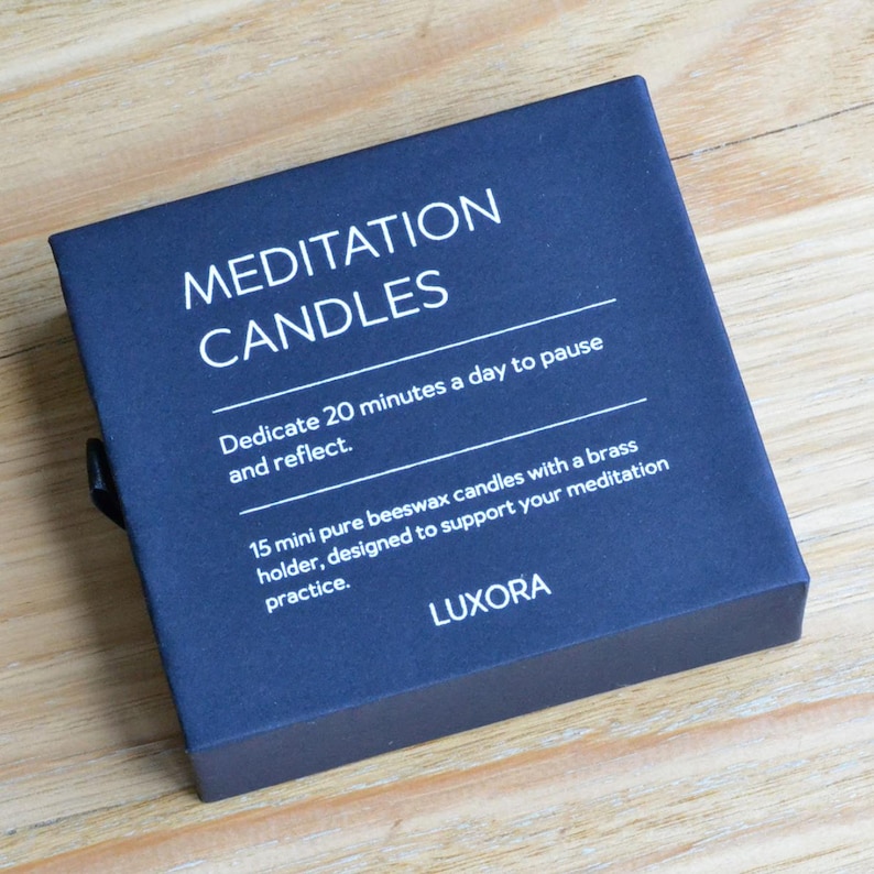 May include: A navy blue box of meditation candles. The box reads "MEDITATION CANDLES" and includes text about dedicating 20 minutes a day to pause and reflect. It also mentions 15 mini beeswax candles with a brass holder.