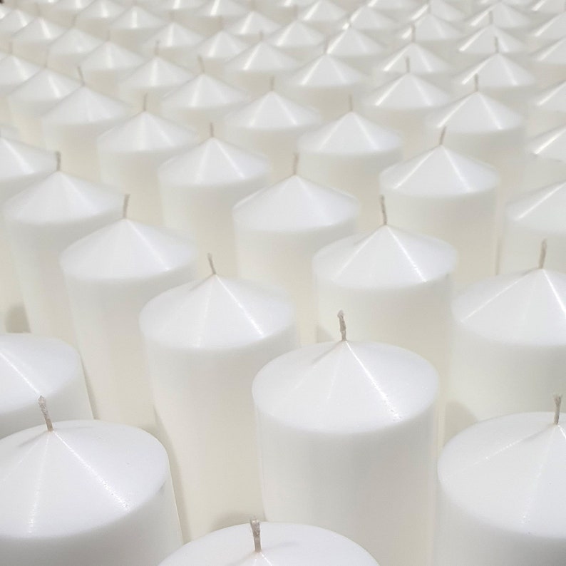May include: A close-up view of a large quantity of white pillar candles arranged in rows. The candles are all the same size and shape, and they have a smooth, polished finish.