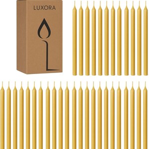 May include: A collection of slender, yellow taper candles arranged around a brown cardboard box. The box is labeled "LUXORA" and features a black candle flame logo. The candles are approximately 8 inches long, and are a warm, golden yellow color.