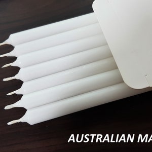 Puede incluir: Un paquete de velas blancas de tipo taper, fabricadas en Australia. Las velas están dispuestas en fila, con las puntas de las velas mirando al espectador. Las velas están en una caja de cartón blanca.