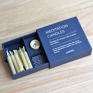 May include: A navy blue box set of meditation candles. The box is open, revealing 15 mini beeswax candles, a brass candle holder, and text that reads "MEDITATION CANDLES". The text also says "Dedicate 20 minutes a day to pause and reflect."
