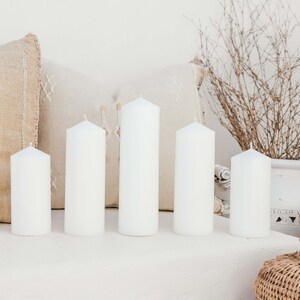 May include: Five white pillar candles of varying heights arranged on a white surface. The candles are in front of a beige and white patterned pillow.
