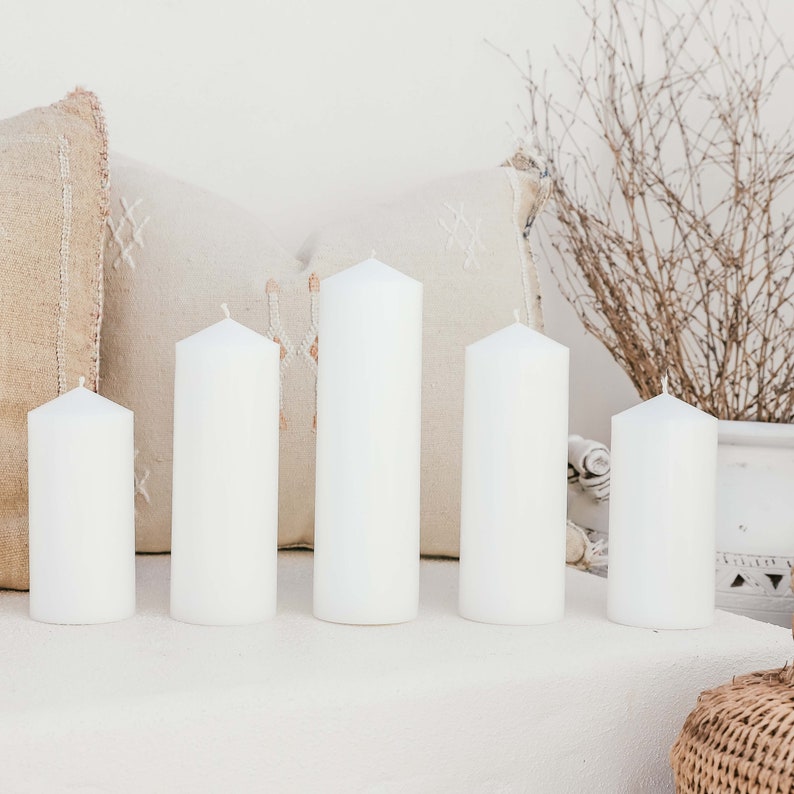 May include: Five white pillar candles of varying heights arranged on a white surface. The candles are in front of a beige and white patterned pillow.