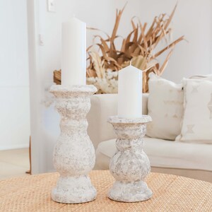 May include: Two white candles in gray textured candle holders on a woven table runner. The candle holders are in a classic design with a round base and a tall, slender stem.