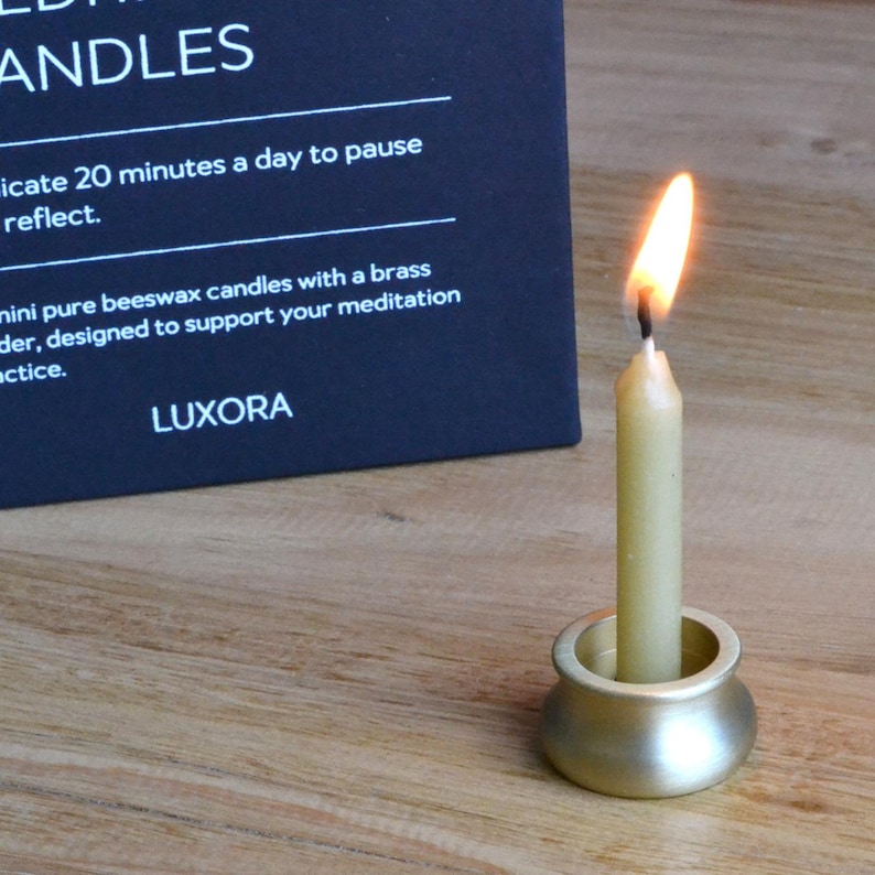 May include: A lit, pale yellow beeswax candle in a brass holder. The candle is burning with a bright orange flame. The text on the black card reads "LUXORA" and describes the candles as designed to support meditation.