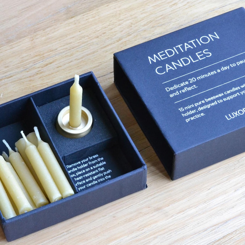 May include: A navy blue box set of meditation candles. The box is open, revealing several small, cream-colored candles and a brass candle holder. The box reads "MEDITATION CANDLES" and includes instructions.