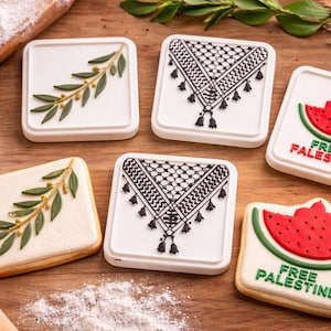 May include: Square cookies with white icing and various designs. Some feature olive branches, others a black and white keffiyeh pattern, and some have a watermelon slice with the text "FREE PALESTINE" in green.
