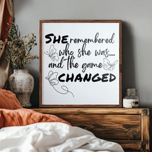 May include: A framed white print with the text "SHE remembered who she was... and the game CHANGED" in black. The print features butterfly illustrations. The frame is brown, and the print is displayed on a wooden nightstand.