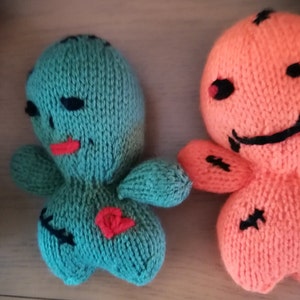 May include: Two small, hand-knitted dolls, one teal and one bright orange. The teal doll features black stitched eyes, a red mouth, and a red heart patch. The orange doll has black stitched eyes and a wide black stitched mouth, with black cross-stitch details.
