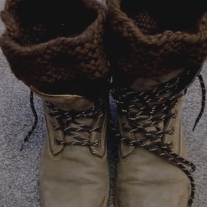 Puede incluir: Un par de botas de cuero marrón claro con puños de punto marrón oscuro en la parte superior. Las botas tienen una punta redondeada, costuras amarillas a lo largo de los bordes y cordones moteados en blanco y negro a través de ojales metálicos.