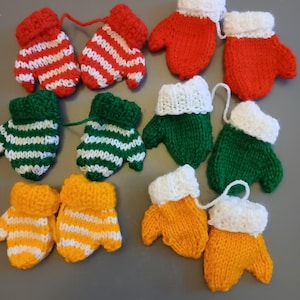 Op de afbeelding: Een verzameling kleine, gebreide wanten in verschillende kleuren, waaronder rood, groen en geel. Sommige wanten hebben witte strepen, andere zijn effen. Elke want heeft een witte manchet en sommige hebben een touwtje.