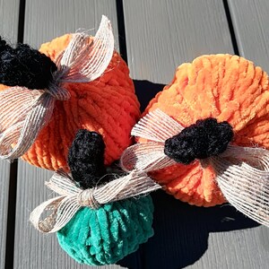 May include: Two orange and one green plush pumpkin decorations made of soft, ribbed yarn. Each pumpkin features a black crocheted stem and a decorative tan burlap ribbon tied around the base of the stem, creating a textured, rounded appearance.