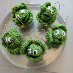 May include: Five green knitted cabbage ornaments with googly eyes and black felt mouths. The ornaments are hanging from gold string on a clear glass plate.