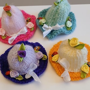 May include: Four small knitted hats in various colours: pink, turquoise, purple, and orange. Each hat is adorned with fabric flowers, bows, and ladybug and bee decorations. These miniature hats have a brim and a rounded crown, suitable for dolls or crafts.