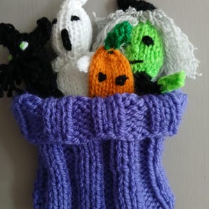 May include: A knitted Halloween-themed decoration featuring a purple bag overflowing with handmade characters. The bag holds a black cat, a white ghost, an orange pumpkin, and a green witch with gray hair. The figures are made of yarn.