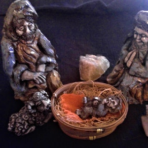 May include: A ceramic nativity scene featuring figures of Mary, Joseph, and baby Jesus in a woven basket. The figures are painted in muted tones of blue, brown, and tan. A sheep is at Joseph's feet. The scene includes two pieces of quartz.