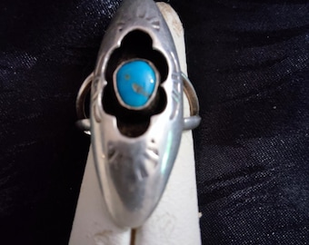 May include: A silver ring with a turquoise stone set in a floral design. The ring has an elongated shape with decorative cutouts and is displayed on a white stand against a dark background.