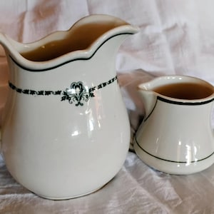 May include: Two white ceramic pitchers. The larger pitcher has a curved handle and a decorative black and green floral band. The smaller pitcher has a rounded body and a small handle. Both pitchers have black trim.
