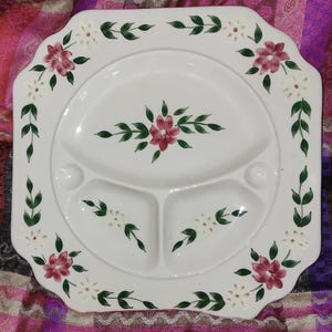 May include: White divided plate with a scalloped edge, decorated with hand-painted floral and leaf designs. The flowers are in shades of pink and white, with green leaves. The plate has three compartments and two small round indentations.
