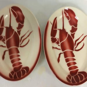 May include: Two oval-shaped plates with a cream-colored background and a dark red lobster design in the center. The plates have a dark red border. The lobster design is detailed with lighter red accents.