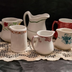 May include: A set of six vintage white ceramic creamer pitchers with various floral and geometric designs. The pitchers are arranged on a white crocheted doily.