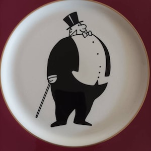 May include: A white ceramic plate with a burgundy rim and a gold trim. The plate features a black and white illustration of a man in a top hat and tuxedo holding a cane.