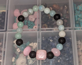 May include: A beaded bracelet featuring a mix of smooth light blue, pale green, and pink beads interspersed with textured black lava stone beads. The design includes a central square pink bead and a variety of round polished stones in pastel tones.
