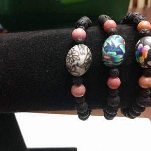 May include: Three beaded bracelets featuring black porous lava stone beads, smooth pink spherical beads, and a single larger focal bead on each. The focal beads display distinct patterns: one white with black floral designs, one blue with multicolored swirls, and one black with colorful floral motifs.