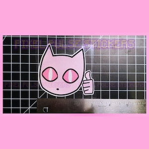 May include: A pink cat sticker with a white outline and a thumbs-up gesture. The cat has large, oval eyes with pink centers. The sticker is on a black grid with a ruler.