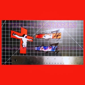May include: Sticker set featuring anime-style eyes and a red cross with a crucified figure. The stickers are on a black grid surface with a ruler for scale. The background is a solid red.