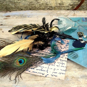 Könnte beinhalten: Eine Auswahl an dekorativen Federn in Gold, Schwarz und Pfauenfedern, zusammen mit einer Pfauenfeder, einem Stift und einem zerbrochenen Glas. Die Gegenstände sind auf einer hellblauen Oberfläche mit handschriftlichen Notizen und einem blauen Umschlag angeordnet.