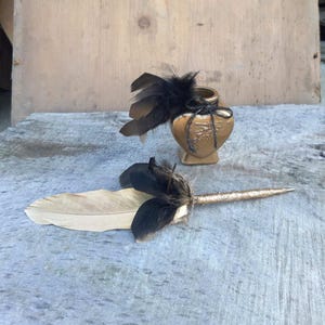 Könnte beinhalten: Ein goldfarbener Tintenfass und ein Federhalter mit schwarzen und beigen Federn. Der Stift hat eine goldglitzernde Spitze. Das Tintenfass ist herzförmig mit einer schwarzen Schleife und Federakzenten. Die Artikel befinden sich auf einer grauen Oberfläche.