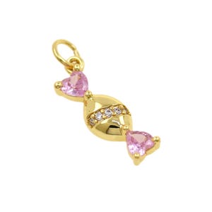 May include: A gold-colored charm in the shape of a wrapped candy. The charm features pink heart-shaped gems at each end and a band of clear gems in the center. The charm has a small loop at the top for attachment.