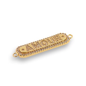 May include: Gold-tone charm with the word "AMOUR" in raised letters, surrounded by clear rhinestones. The charm has two jump rings for attaching to a bracelet or necklace.