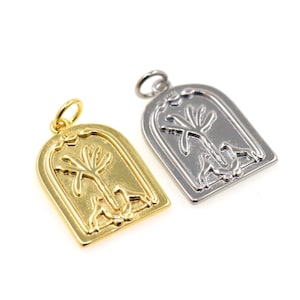 May include: Two metal pendants, one gold-toned and one silver-toned, each featuring a stylized tree design within an arched frame. The pendants have a loop at the top for attaching to a chain or other jewelry component. The background is white.