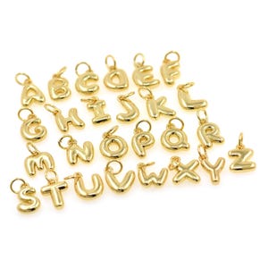 Może przedstawiać: Kolekcja złotych zawieszek z alfabetem, każda z pętelką do mocowania do łańcuszka lub bransoletki. Litery są w zaokrąglonej, bąbelkowej czcionce i są wyświetlane na białym tle.